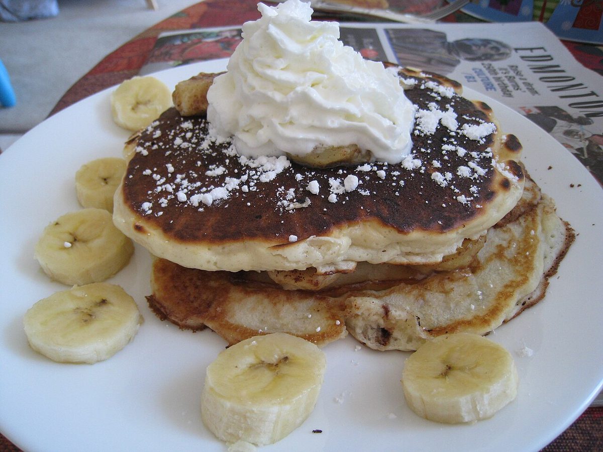 Stos pancakes z bitą śmietaną, plasterkami banana i cukrem pudrem na białym talerzu